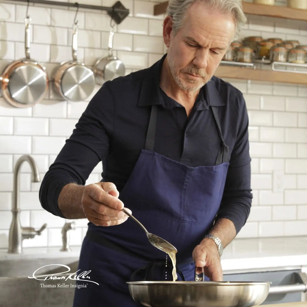 A man in a navy apron pours liquid from a spoon into a Thomas Keller Commercial Clad Stainless Steel pan from the 10-Piece Cookware Set, in a modern kitchen with white subway tiles and shelves of spices.
