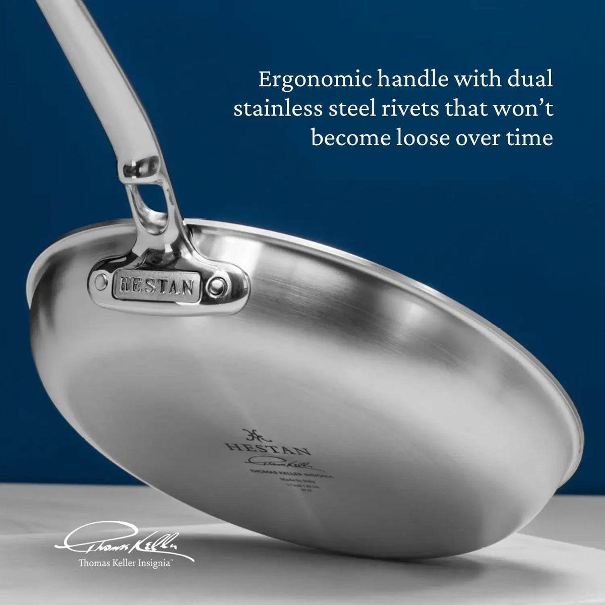 A Thomas Keller Commercial Clad stainless steel frying pan from the 10-Piece Cookware Set is shown on a blue background. Its ergonomic handle, dual rivets, and Tri-Ply Stainless Steel design ensure durability, with Hestan branding and Keller’s signature featured.