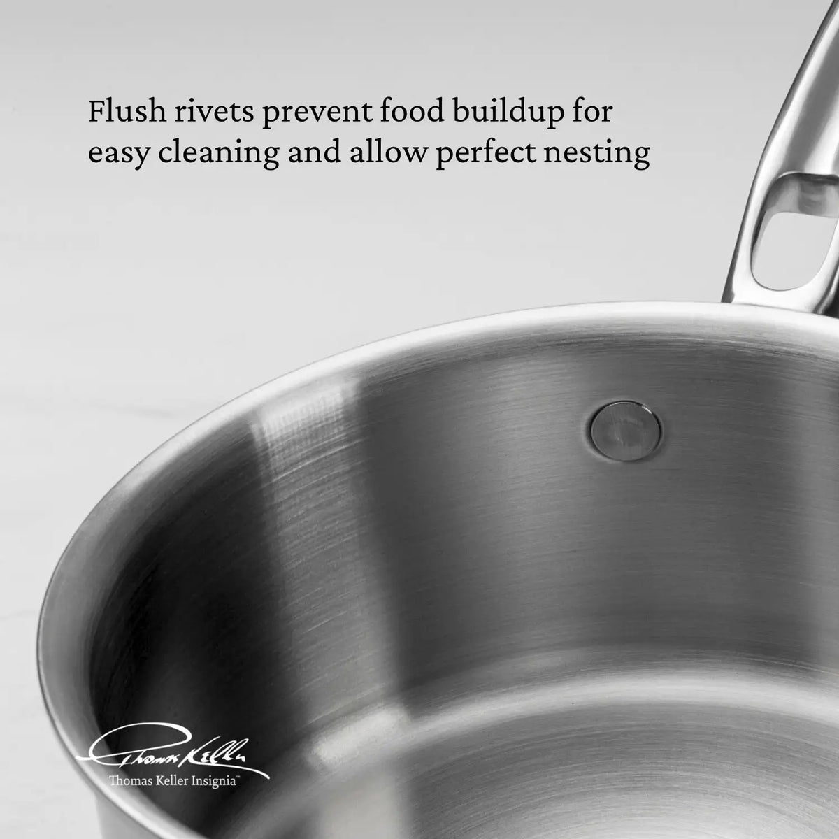 A close-up of a Thomas Keller Commercial Clad Stainless Steel 10-Piece Cookware Set pan shows flush rivets that prevent food buildup for easy cleaning and perfect nesting. The pan features a handle and a seamless tri-ply stainless steel interior.
