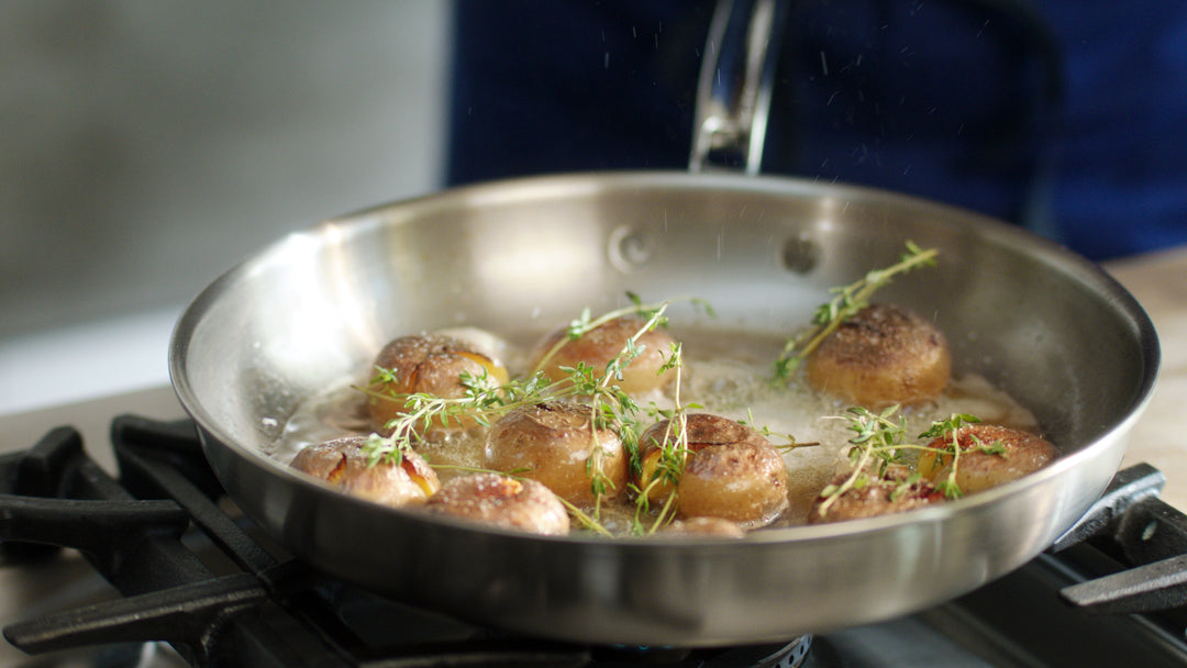 A stainless steel pan on a stovetop contains partially cooked potatoes with browned tops, garnished with fresh thyme sprigs. The pan is bubbling with oil or butter.