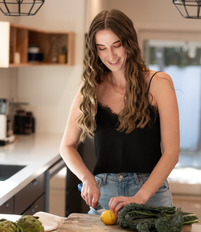 A woman in a black tank top and jeans is chopping a lemon in a kitchen. She stands at a wooden countertop with leafy greens nearby. The kitchen has modern decor with a window in the background.