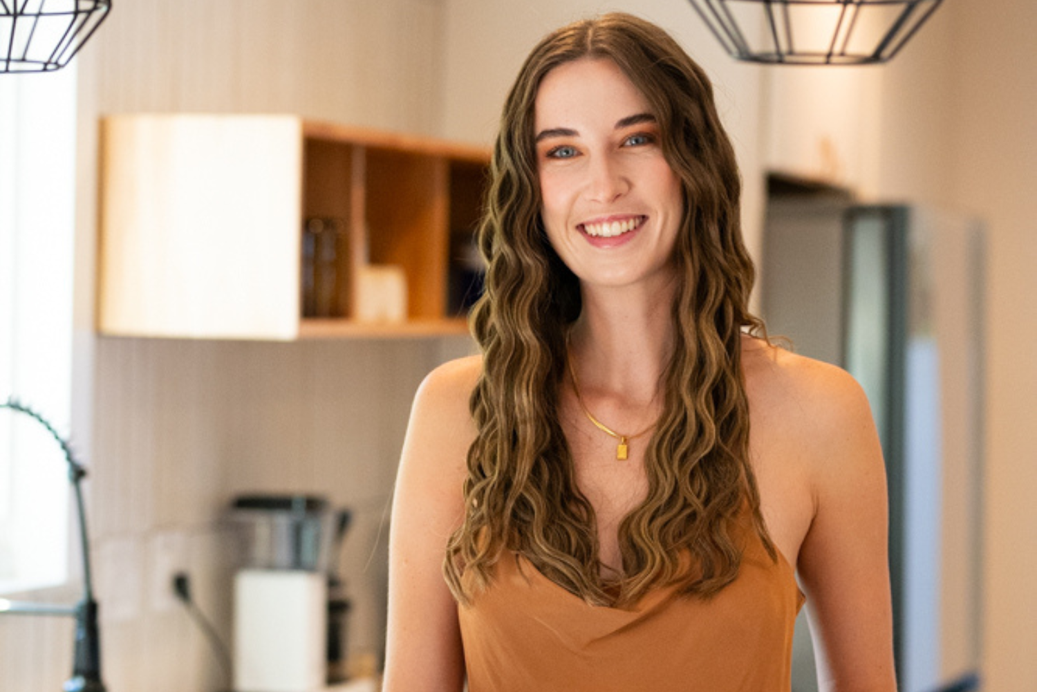 A woman with long wavy hair smiles while standing in a modern kitchen. She is wearing a sleeveless brown top and a gold necklace. In the background, there are shelves, a sink, and a refrigerator visible.