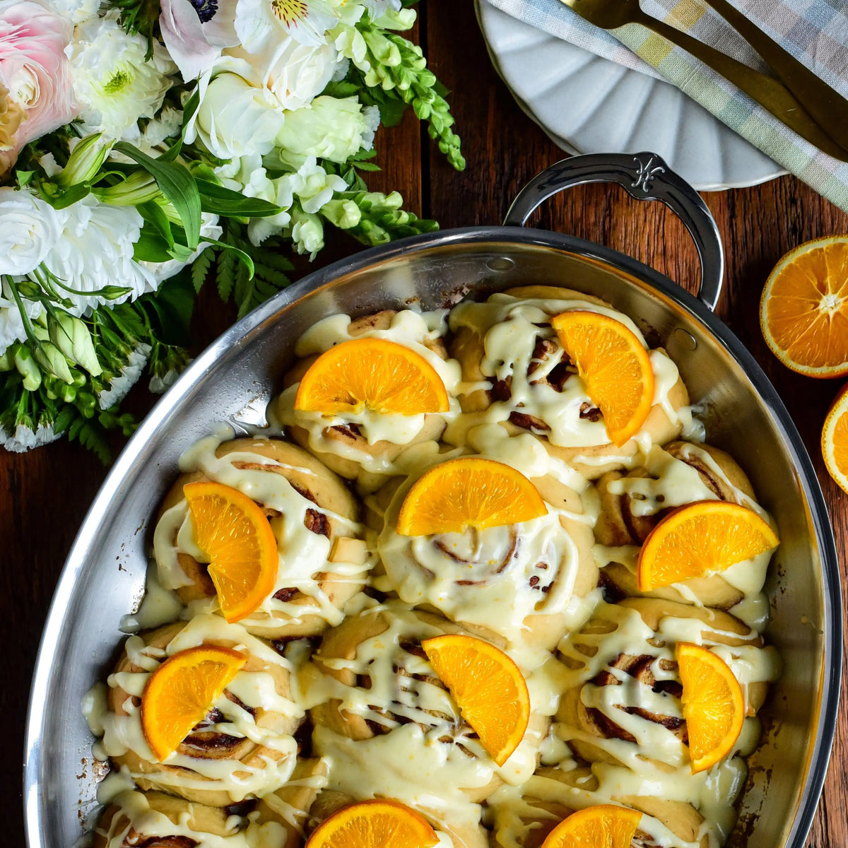 A Hestan Provisions 17.5-Inch x 14-Inch Clad Oval Roaster holds frosted cinnamon rolls garnished with orange slices, surrounded by sliced oranges, a plate with cutlery, and white flowers on a wooden table for an elegant oven-to-table display.