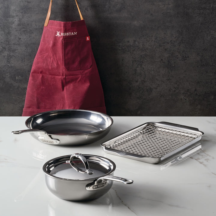 Against a dark backdrop stands the Titanium Grilling Essentials Cookware Set by NanoBond. In front, the 6-piece collection—which includes a frying pan, saucepan with glass lid, and roasting pan—is arranged on a white countertop.