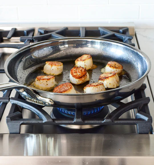 Scallops are searing in a stainless steel frying pan on a lit gas stove. The scallops are browned and sizzling in melted butter or oil. The background features a white tiled backsplash.