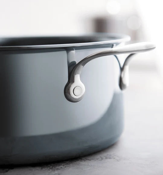Close-up of a shiny gray cooking pot with a silver handle, resting on a smooth surface. The pots reflective surface captures light, emphasizing its sleek design. The background is softly blurred.