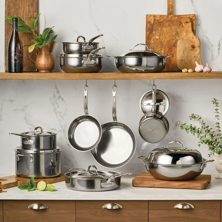 A kitchen features the NanoBond 18-Piece Limited Edition Titanium Cookware Set with Chef's Pan on a wooden shelf and counter, with pots, pans, lids, cutting boards, a bottle, and fresh herbs accenting a marble wall backdrop.