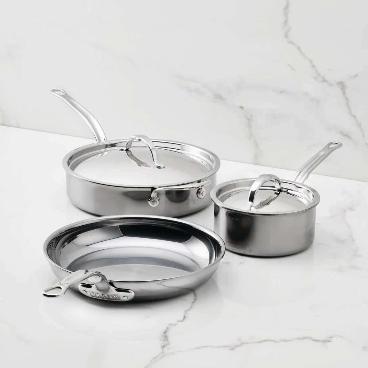 Three pieces from the NanoBond® 5-Piece Titanium Essential Cookware Set are displayed on a marble surface: a frying pan, a sauté pan with lid, and a saucepan with lid. Each item features metal handles and a polished titanium finish, highlighting the durability and quality associated with premium NanoBond® cookware. The background is a white marble wall.