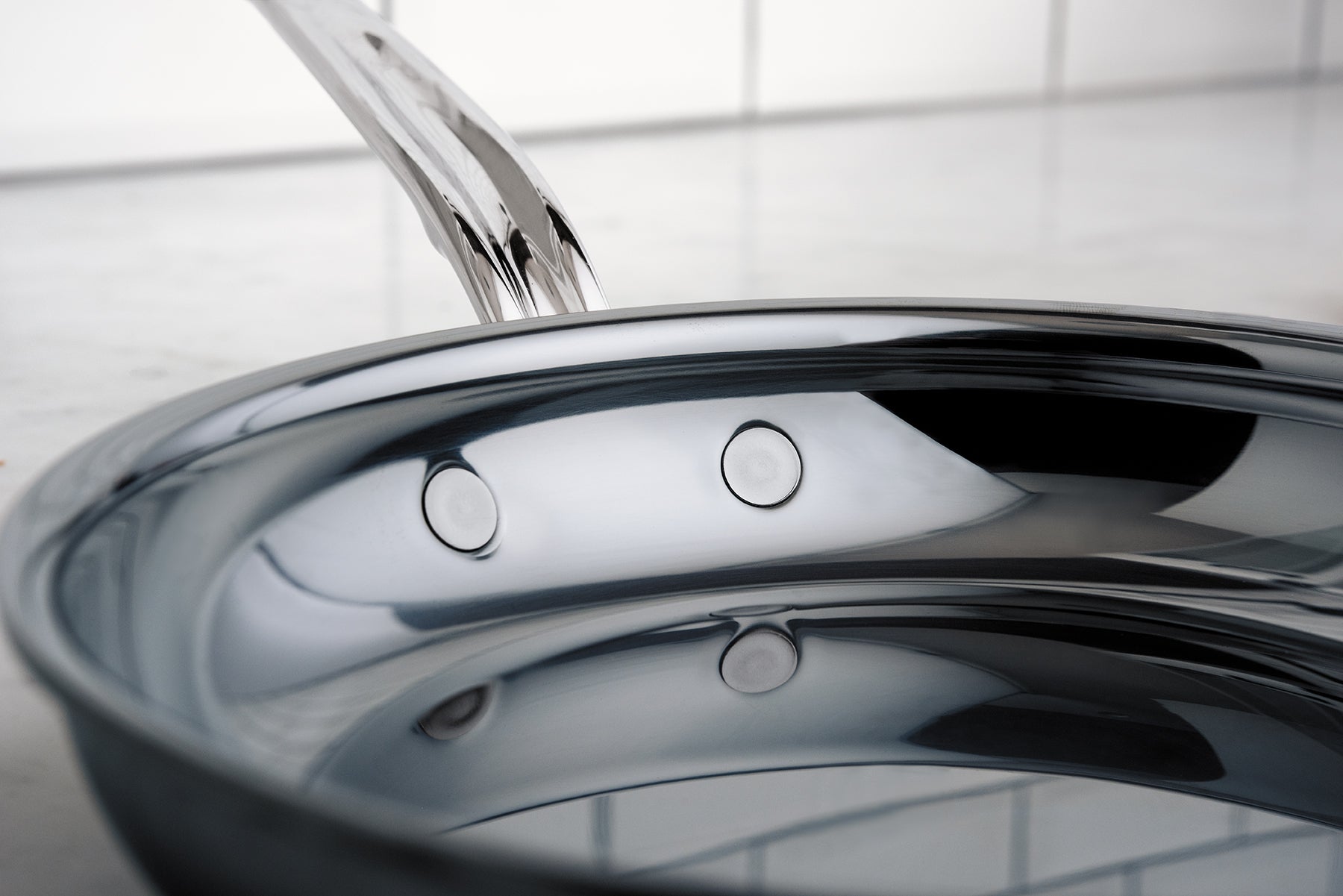 Water is being poured into a shiny stainless steel pan, creating ripples on the surface. The background is a smooth, light-colored surface with tiled walls.