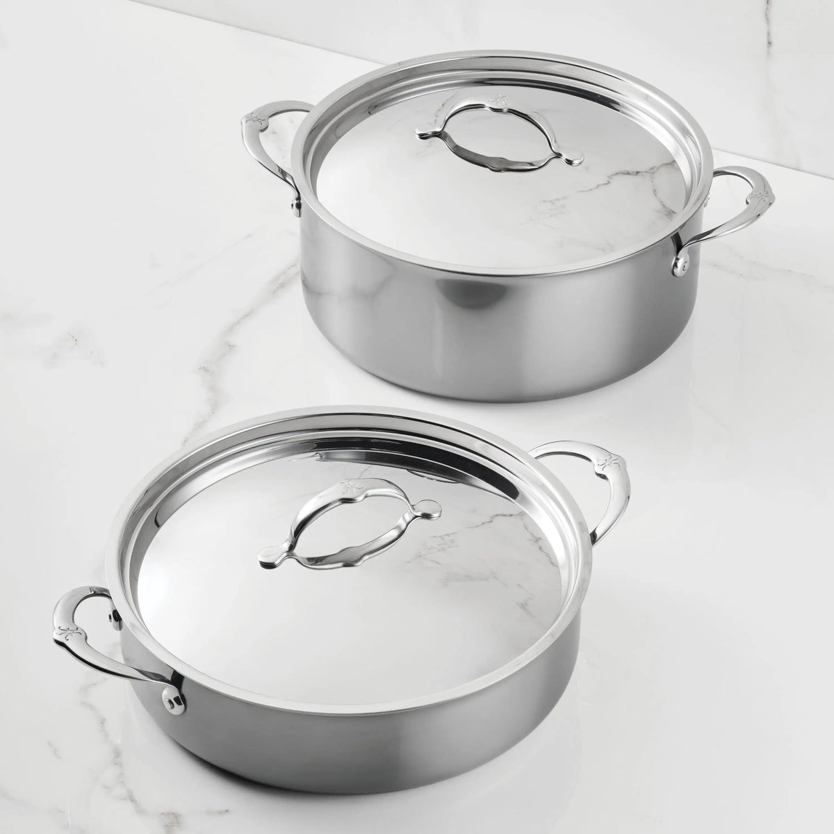 Two NanoBond Titanium Rondeaus with lids and handles sit on a white marble countertop, one slightly behind the other, both highlighting their polished titanium surfaces.
