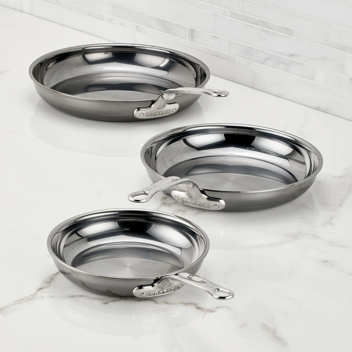 Three NanoBond Titanium Skillets in various sizes are displayed on a white marble countertop with a white tile backsplash, showcasing their shiny exteriors and sleek metal handles.