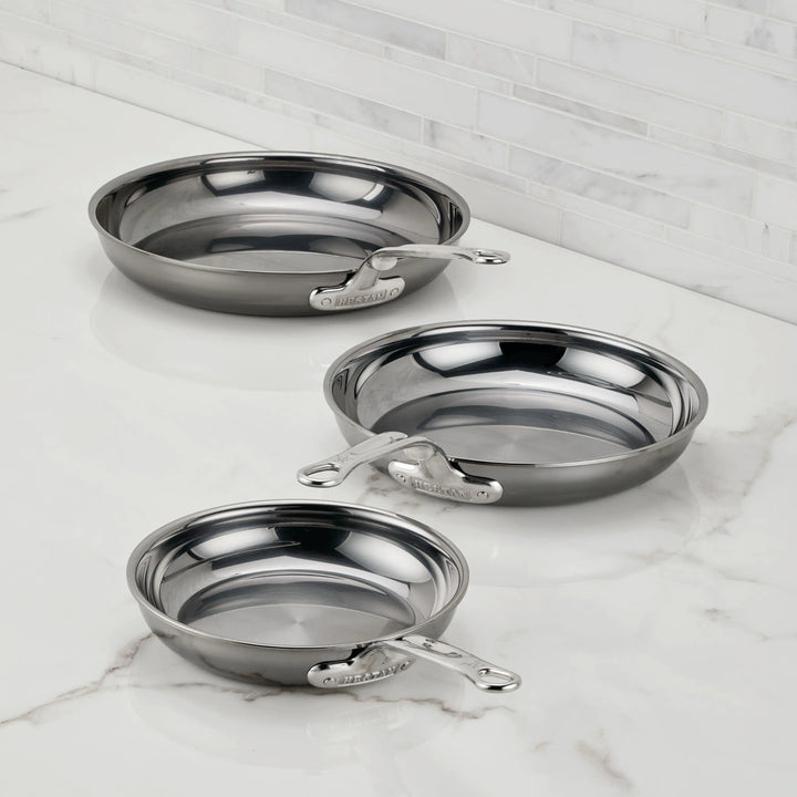 Three NanoBond Titanium Skillets in various sizes are displayed on a white marble countertop with a tiled backsplash. Each frying pan showcases a sleek metallic handle and non-reactive titanium construction.