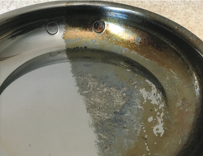 A metal pan showing a distinct line between a clean, shiny half and a dull, discolored half. The clean side reflects light, while the discolored side appears tarnished with visible patches of residue.