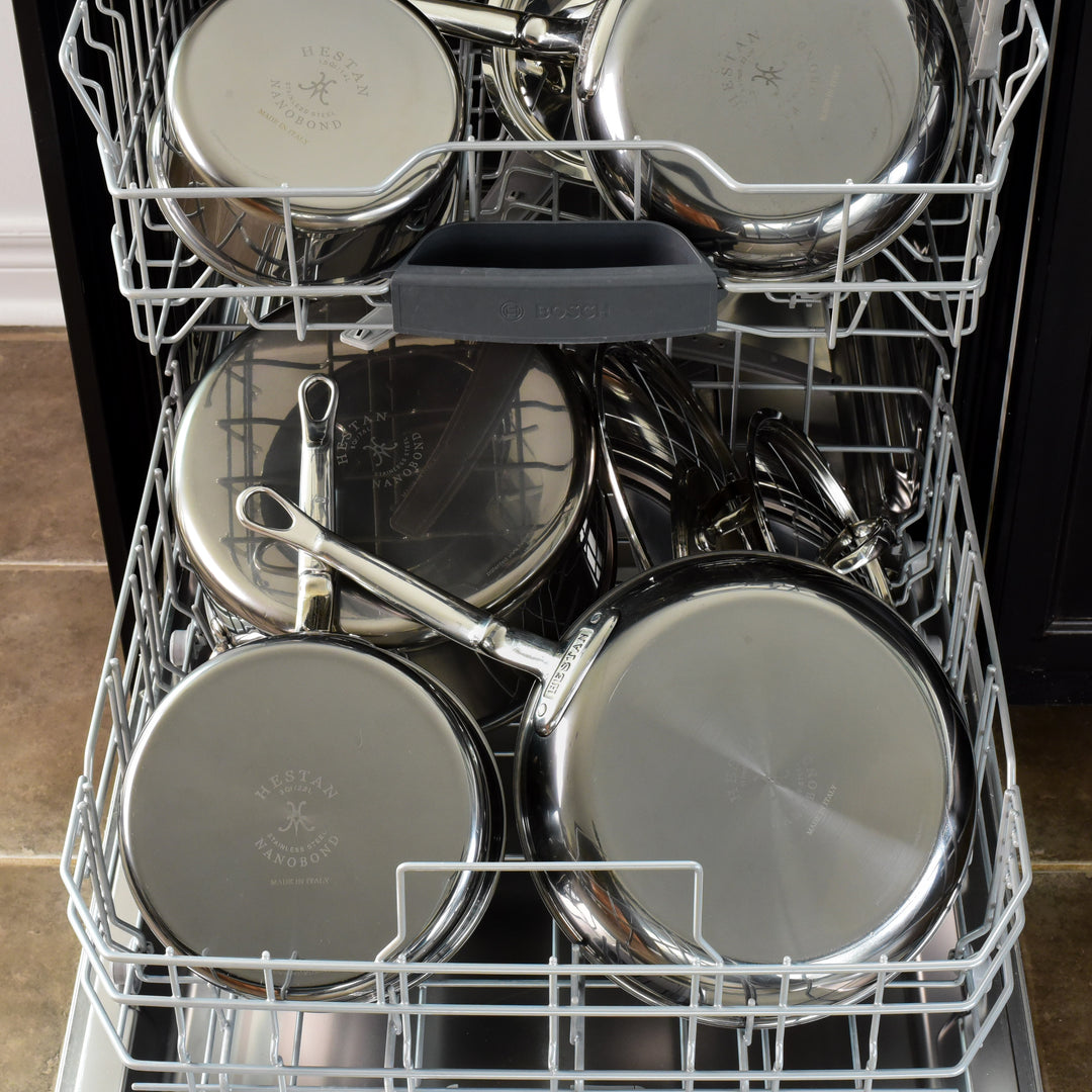 Open dishwasher loaded with various stainless steel pots, pans, and lids. The cookware is neatly arranged on multiple racks, reflecting ambient light. The interior is clean and the door part is visible at the bottom.
