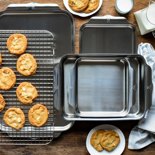 Baking scene with stainless steel pans of various sizes on a wooden surface. Freshly baked cookies are on a cooling rack and in a small plate. Two glasses of milk are nearby, along with a folded striped cloth.
