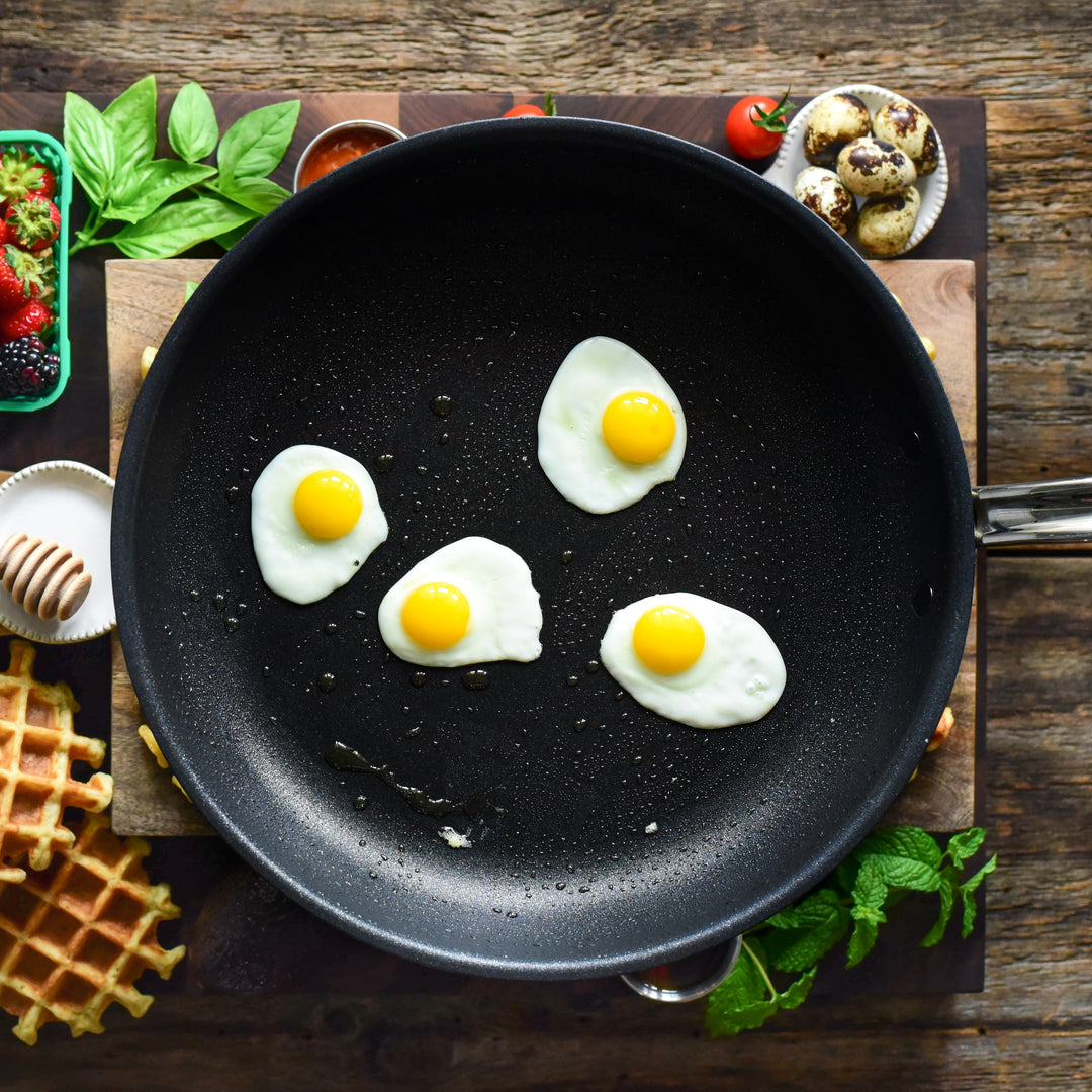 A frying pan with four small sunny-side-up eggs is on a wooden surface. Surrounding the pan are waffles, a bowl of strawberries, herbs, quail eggs, cherry tomatoes, and a small dish of honey, creating a breakfast-themed arrangement.