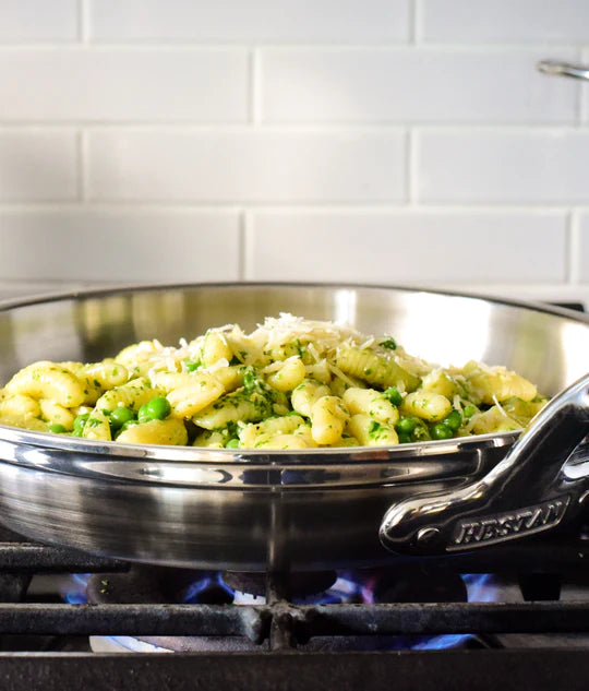 A stainless steel pan on a gas stove contains cooked gnocchi mixed with green peas and topped with grated cheese. The stove has a visible blue flame. White subway tiles are in the background.