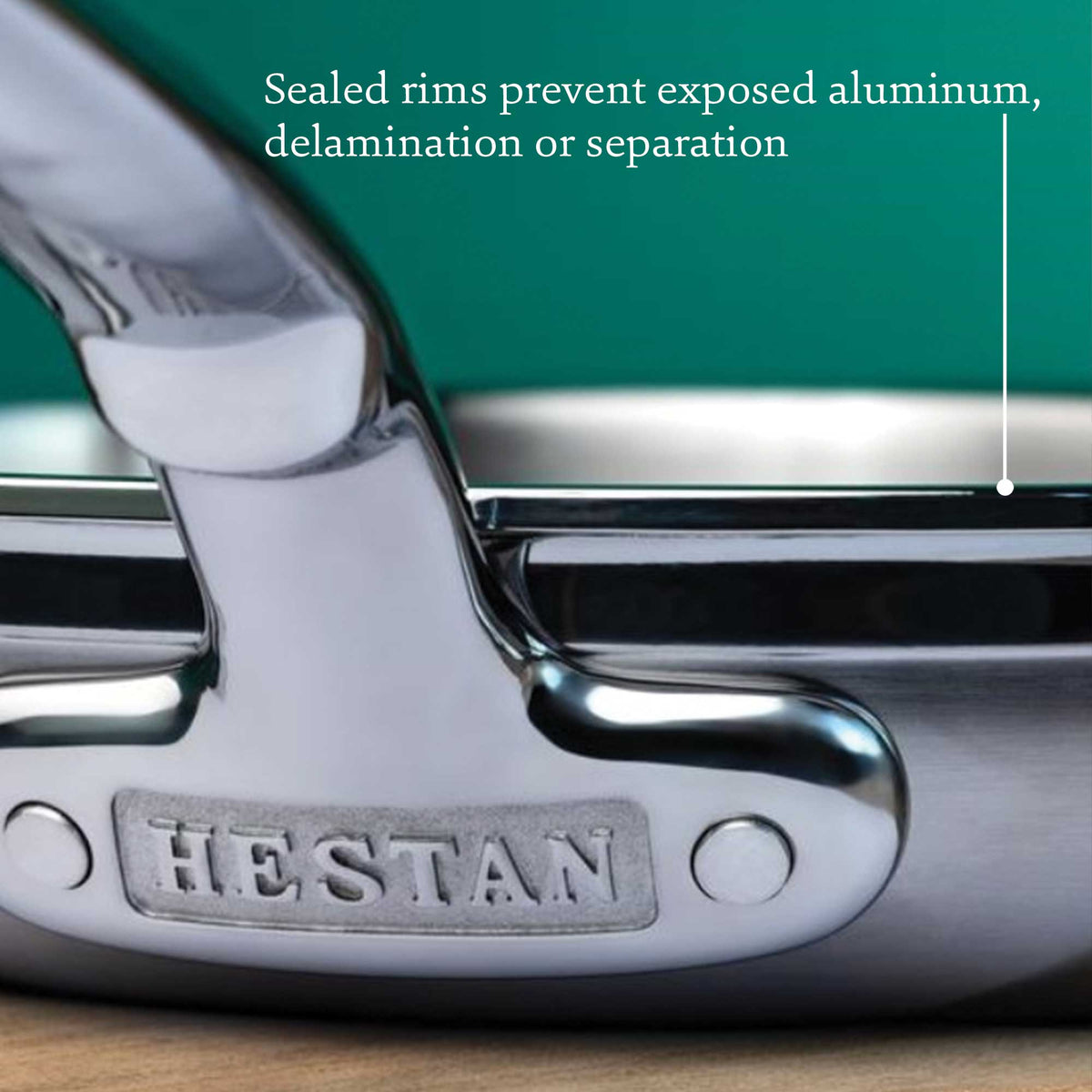 Close-up of a ProBond Professional Clad Stainless Steel Essential Pan, highlighting the handle with HESTAN engraved. The focus is on the riveted joint and tri-ply aluminum core, promising durability with sealed rims to prevent aluminum exposure or delamination.