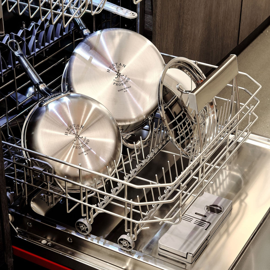 Stainless steel frying pans and lids are neatly arranged in the bottom rack of an open dishwasher. The dishwasher is located in a modern kitchen setting with wooden cabinets in the background.