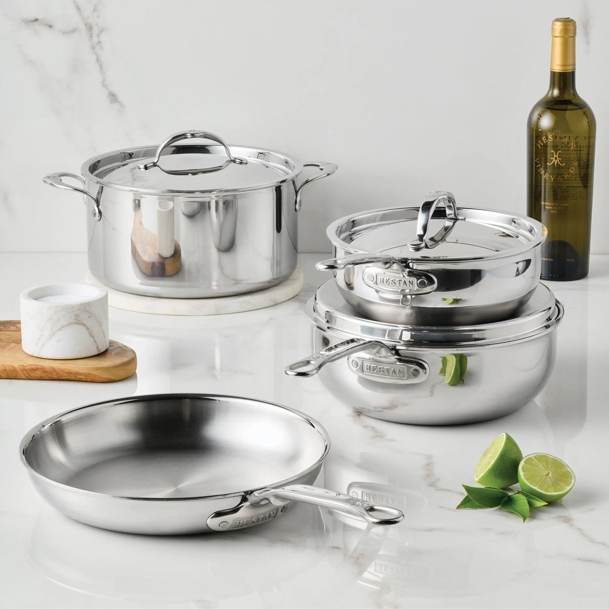 The ProBond® Luxe™ Polished Clad Stainless Steel Signature Set, 7-Piece, is displayed on a marble countertop alongside a marble container, white wine, and cut limes.