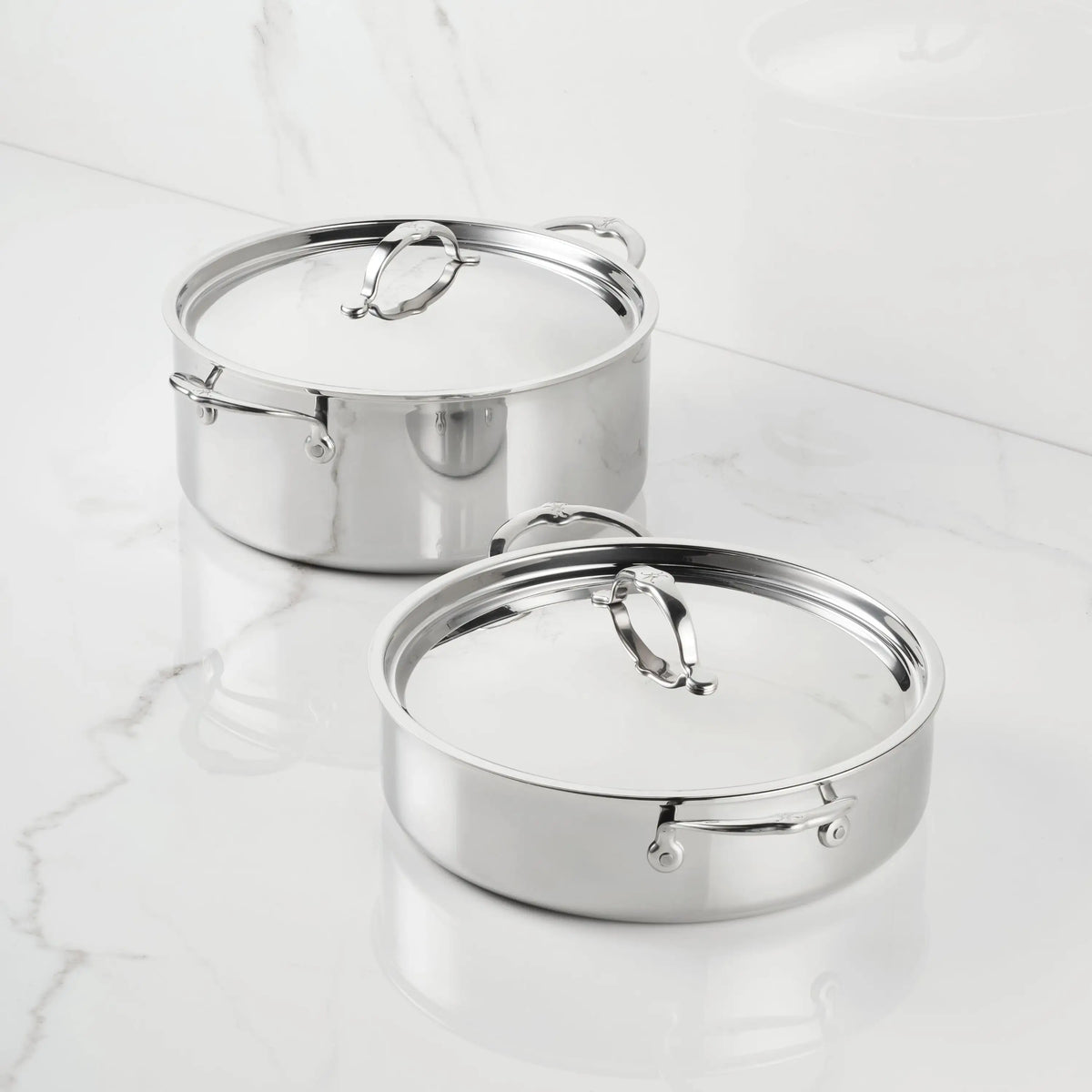 Two ProBond® Luxe™ Polished Clad Stainless Steel Rondeaus with lids sit on a white marble countertop, displaying their polished surfaces, sturdy handles, and matching metal lids that highlight premium cookware design.