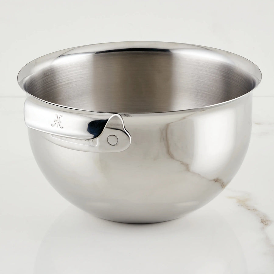 A Hestan Provisions 7-Quart Stainless Steel Mixing Bowl with a rolled rim and side handle sits on white marble. Dishwasher safe, it has a reflective finish and an engraved letter K on the handle.
