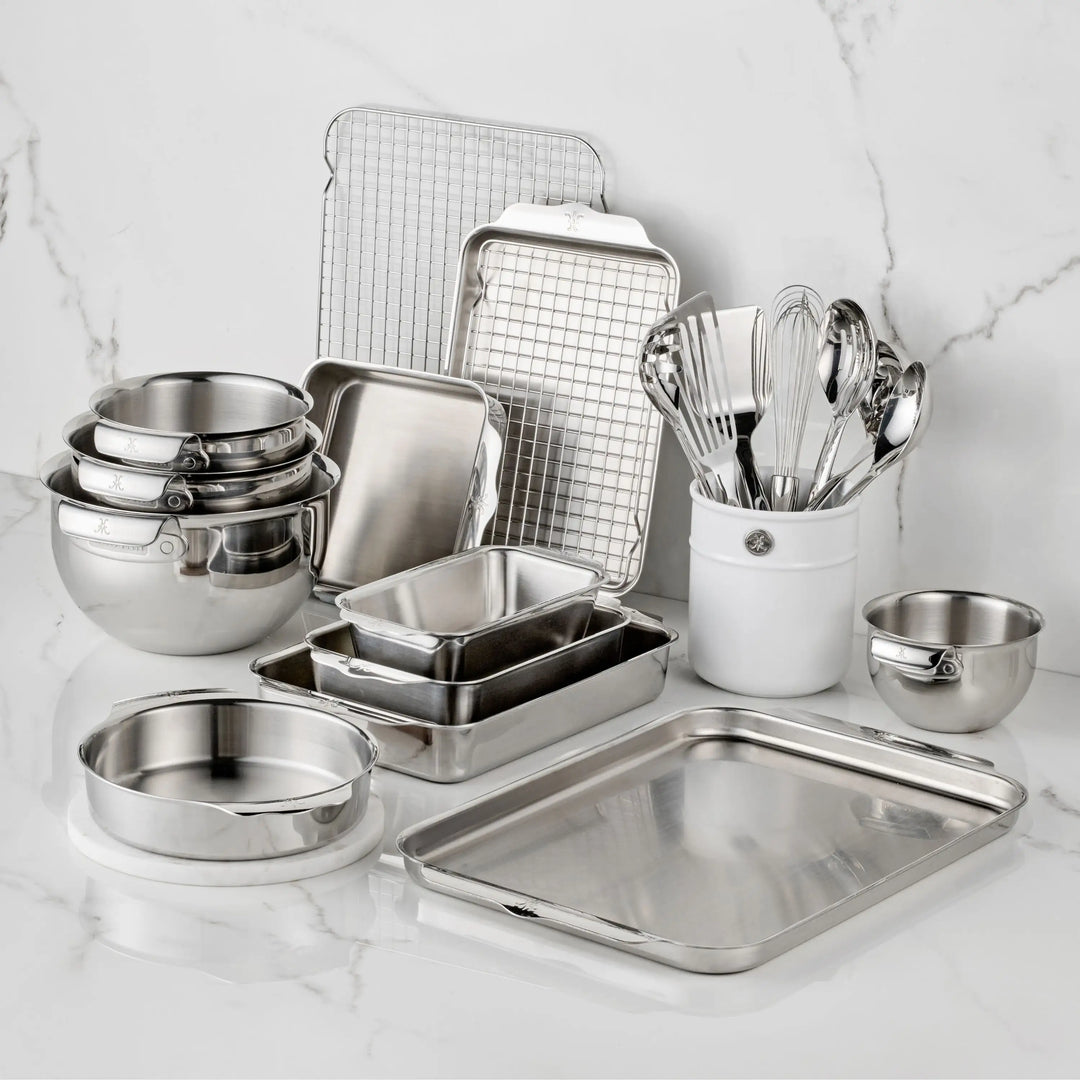 The Hestan Provisions OvenBond Tri-ply Epicurean Stainless Steel 21-Piece Set, featuring bakeware, mixing bowls, pans, a cooling rack, measuring cups, and utensils in a ceramic holder, is arranged on a white marble countertop.