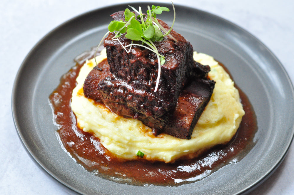 A dish with braised short ribs on a bed of creamy mashed potatoes, topped with microgreens. The meat is glazed with a brown sauce, and the presentation is on a dark plate.