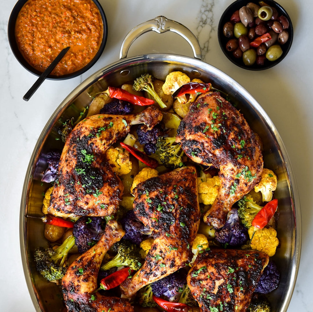 A roasted chicken dish in a large pan with colorful vegetables like cauliflower, broccoli, and peppers. Next to it are a bowl of red sauce with a spoon and a small bowl of assorted olives. The dishes are placed on a light surface.