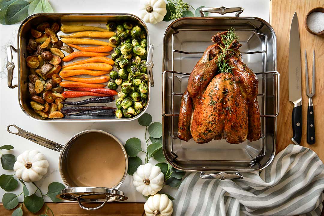 A roasted turkey garnished with herbs is in a pan on the right. The left pan contains roasted vegetables: carrots, brussels sprouts, and potatoes. A saucepan of gravy, a striped cloth, knives, and small white pumpkins are nearby.