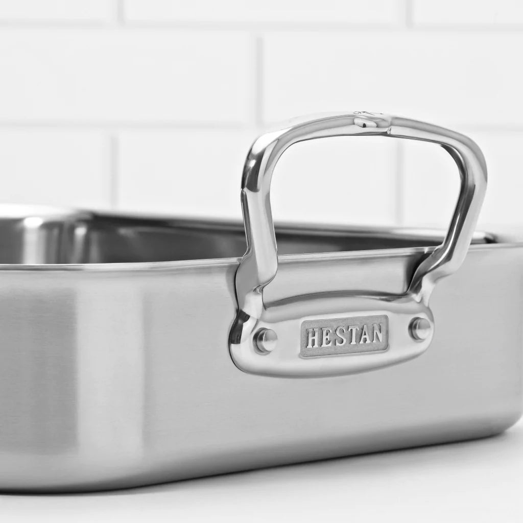 Close-up of a stainless steel roasting pan handle, attached to the pan with visible screws. The brand name HESTAN is embossed on a metal plate below the handle. The background features white subway tiles.