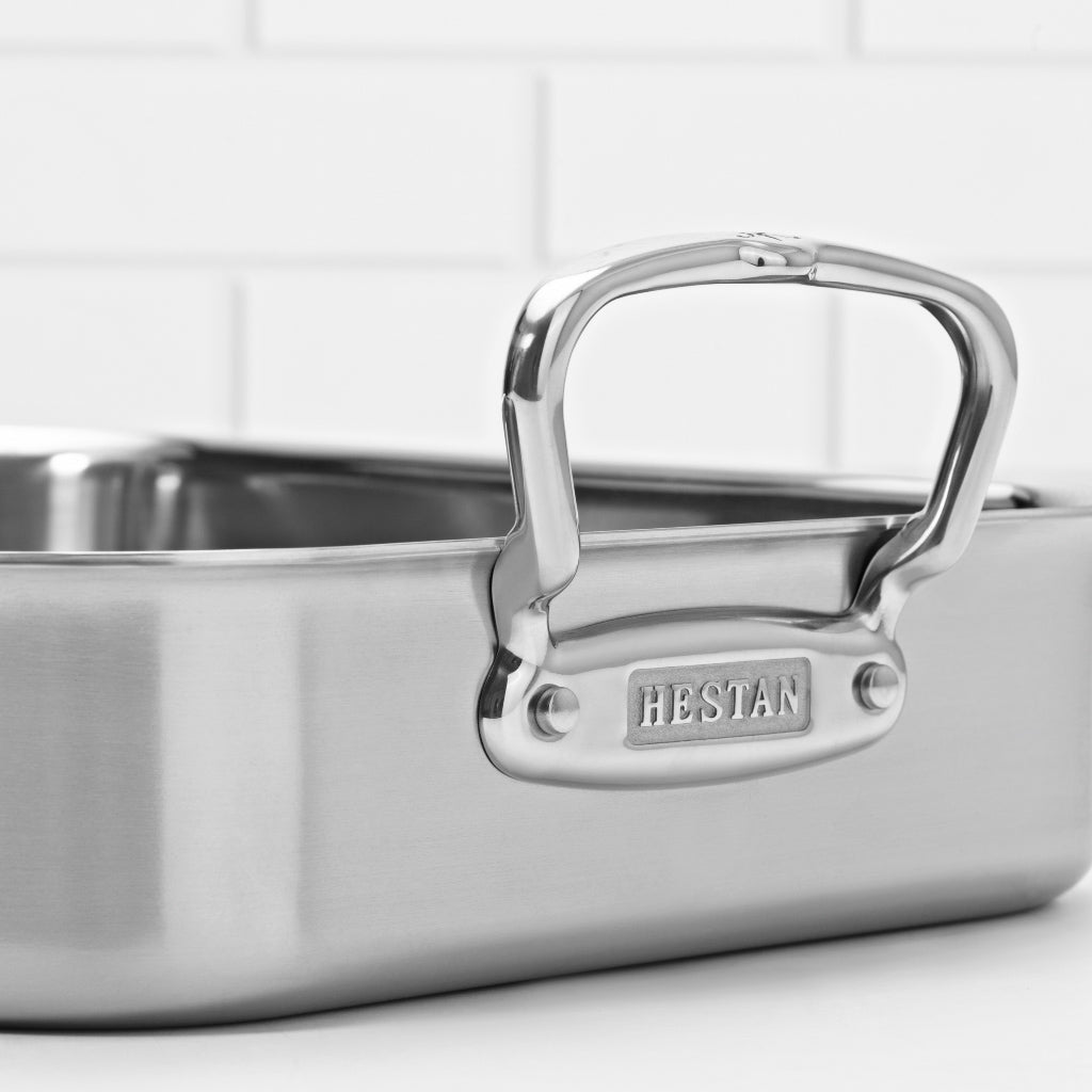 Close-up of a stainless steel baking pan with a large handle engraved with HESTAN. The handle is riveted to the pan. The background is a partially visible white tile wall.