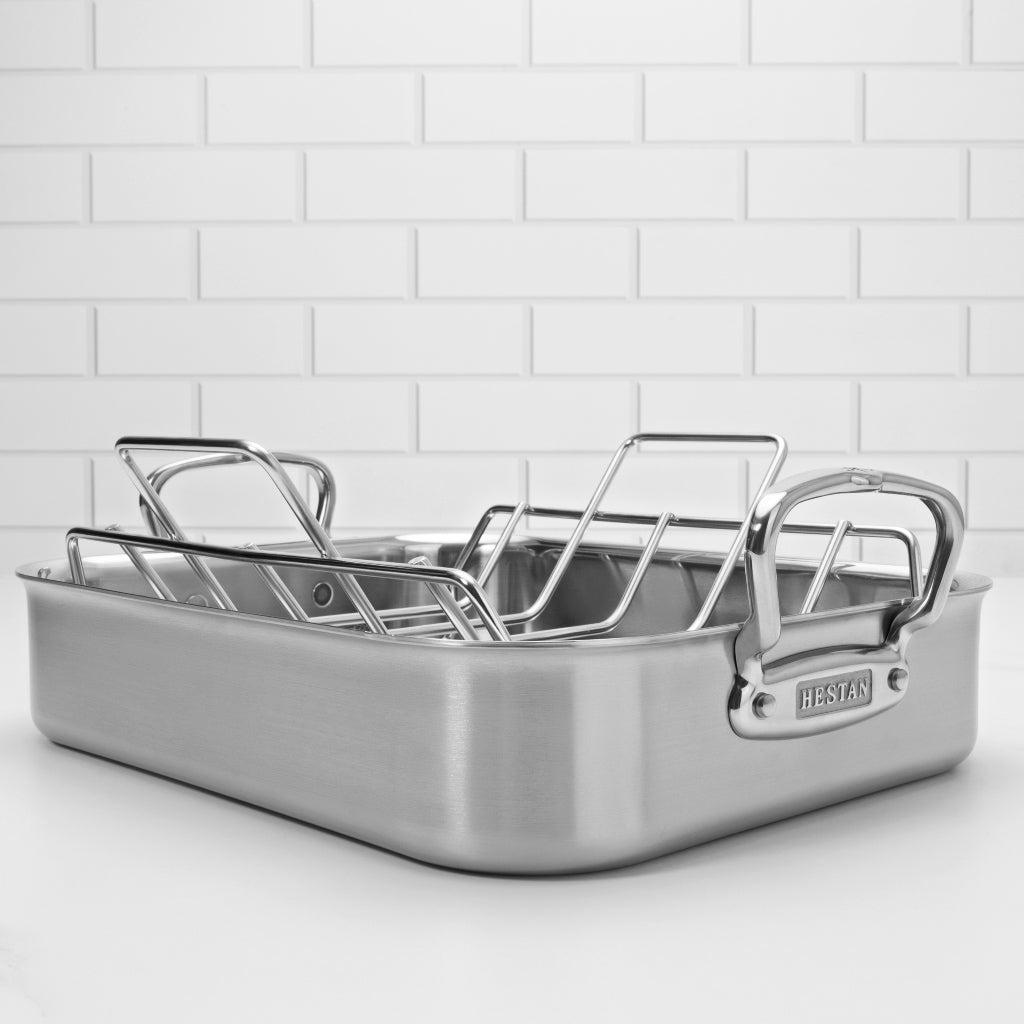A stainless steel roasting pan with a high rack is set on a white countertop. The pan has a rectangular shape with sturdy handles and a polished finish, and is positioned against a white tile backsplash.