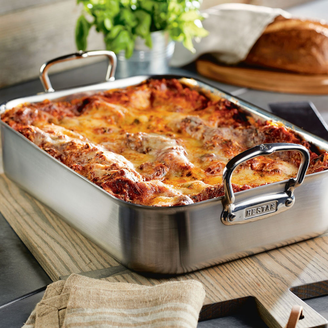 A browned cheese-topped lasagna sits in a NanoBond 28-Piece Titanium Excellence rectangular dish with handles on a wooden cutting board. In the background are a loaf of bread and leafy greens, with a cloth napkin completing this Limited Edition Cookware scene.