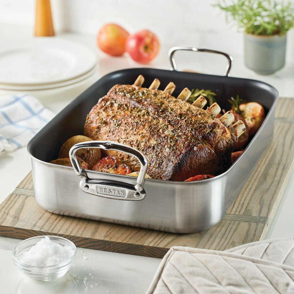 A rack of seasoned beef sits on veggies in a Hestan Provisions Nonstick Clad Stainless Steel Roaster with Rack on a wooden board, with salt, an oven mitt, plates, and apples nearby.