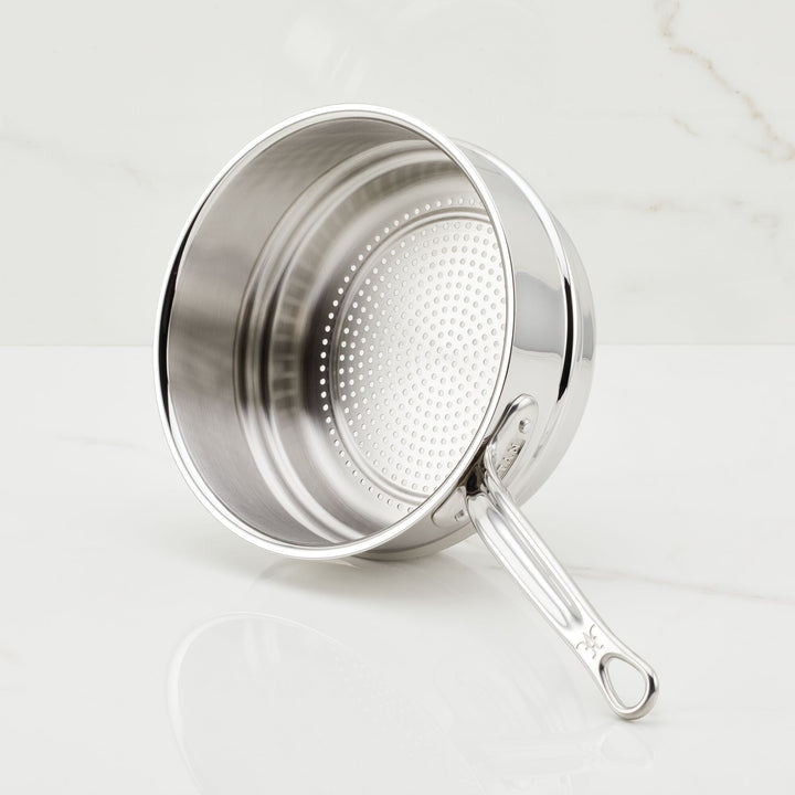 The Hestan Provisions Stainless Steel Steamer Insert, 3-Quart, sits on a white marble surface. It has a perforated base, rivet-mounted handle with a hanging loop, and a reflective interior/exterior for durability and style.
