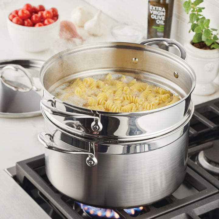 An 8-Quart Stainless Steel Pasta Insert from Hestan Provisions bubbles on the stove, boiling fusilli to perfection. Nearby, olive oil, ripe tomatoes, and a leafy plant adorn the white countertop, setting the scene for an exquisite meal.