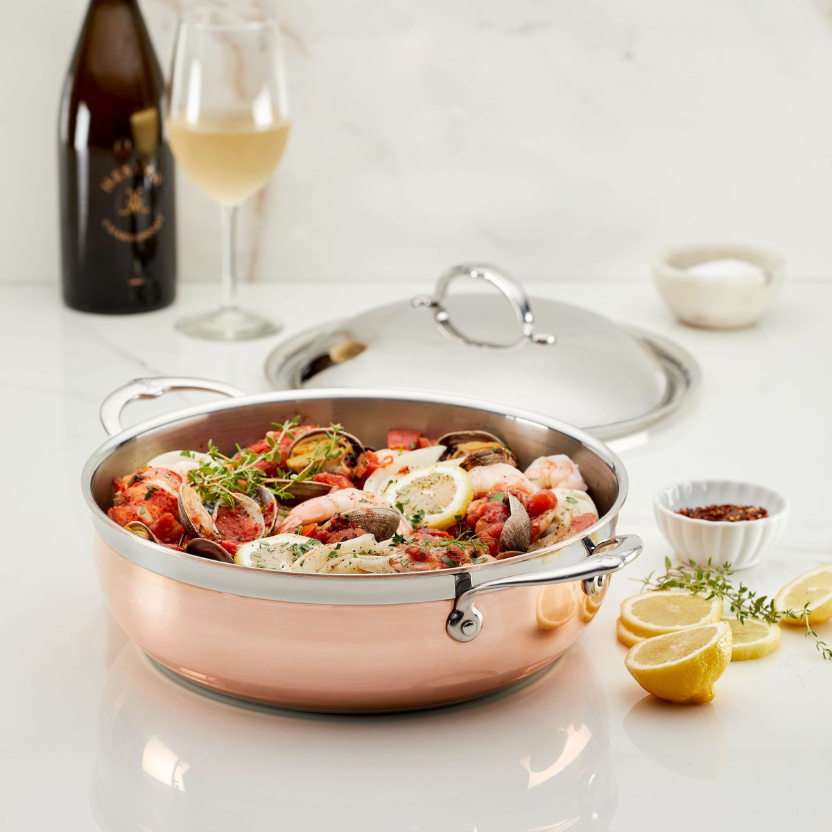 A CopperBond Copper Induction Dutch Oven, 5-quart, filled with a seafood dish featuring shrimp and garnished with lemon slices and herbs, sits on a white table. Nearby are a glass of white wine, a wine bottle, red pepper flakes in a small bowl, a serving lid, and lemon wedges.