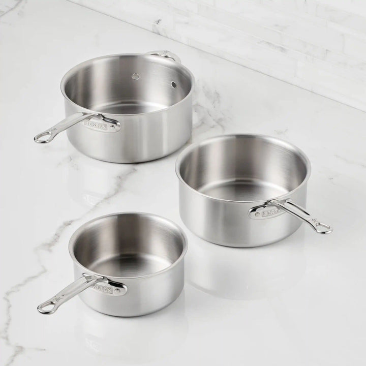 Three Thomas Keller Commercial Clad stainless steel saucepans from the 11-Piece Cookware Set, each with metal handles, are displayed on a white marble countertop against a backdrop of white tiled wall, emphasizing their sleek, professional style.
