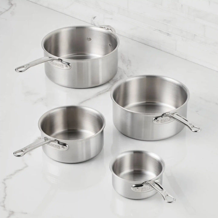 A Thomas Keller Commercial Clad Stainless Steel 0.75-Quart Butter Warmer with a metal handle sits empty on a white marble countertop, set against a white subway-tiled wall.