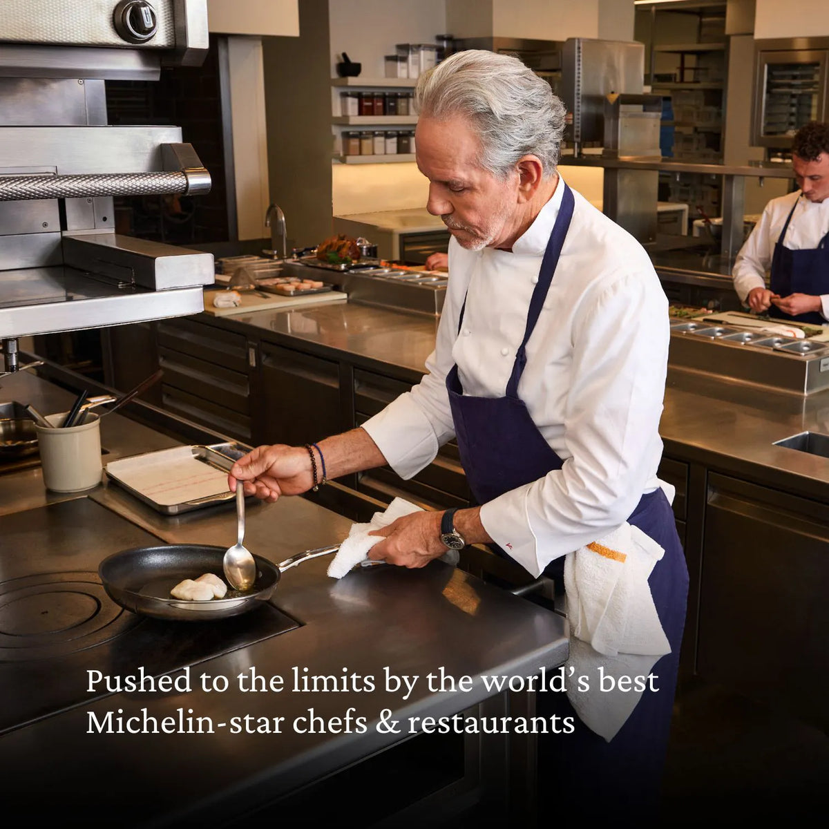 A chef in a white coat and navy apron cooks with the Thomas Keller Commercial Clad Stainless Steel 11-Piece Cookware Set in a pro kitchen. Another chef works behind. Text: Trusted by top Michelin-star chefs & restaurants worldwide.