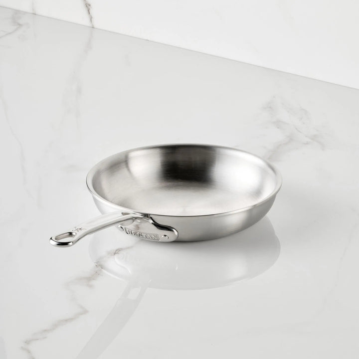 The Thomas Keller Commercial Clad Stainless Steel Skillets feature a chef-designed, long-handled tri-ply construction, shown angled on a white marble countertop with gray veining for a sleek kitchen aesthetic.