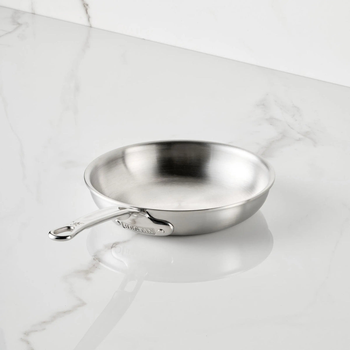 The Thomas Keller Commercial Clad Stainless Steel Skillets feature a chef-designed, long-handled tri-ply construction, shown angled on a white marble countertop with gray veining for a sleek kitchen aesthetic.