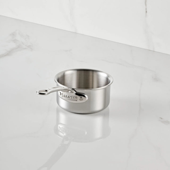 A Thomas Keller Commercial Clad Stainless Steel 0.75-Quart Butter Warmer with a short handle rests on a white marble countertop, featuring the brand name engraved on its handle.