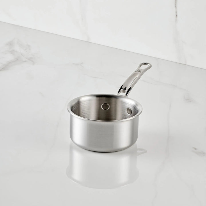 The Thomas Keller Commercial Clad Stainless Steel 0.75-Quart Butter Warmer with a sturdy handle rests on a white marble countertop, reflecting light and elegance amidst matching marble surfaces.