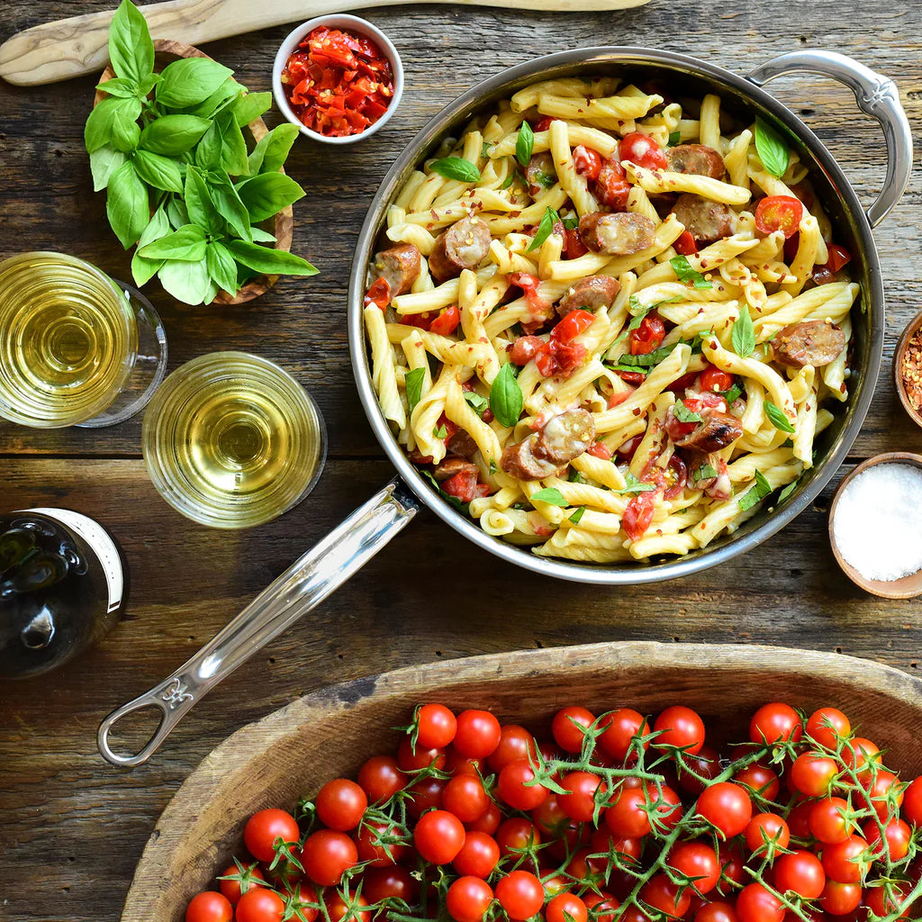 A skillet with pasta, sausage, and tomatoes garnished with basil. Surrounding items include a bowl of cherry tomatoes, two glasses of white wine, a basil bunch, a bottle, and condiments. Everything is set on a rustic wooden table.