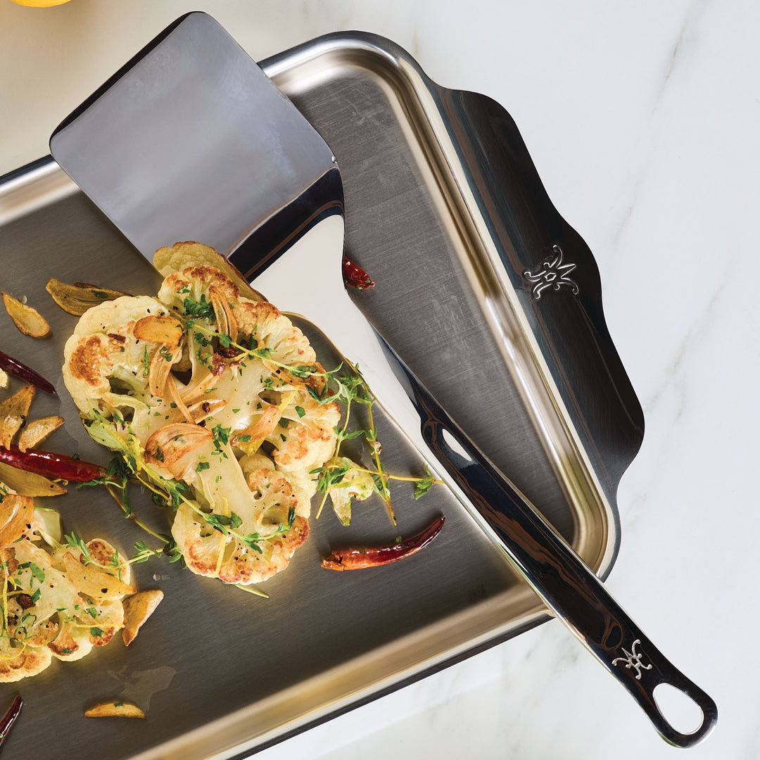 A stainless steel baking tray on a marble surface, featuring roasted cauliflower steaks garnished with herbs and chili peppers. A metal spatula rests partially on the tray, reflecting some light.