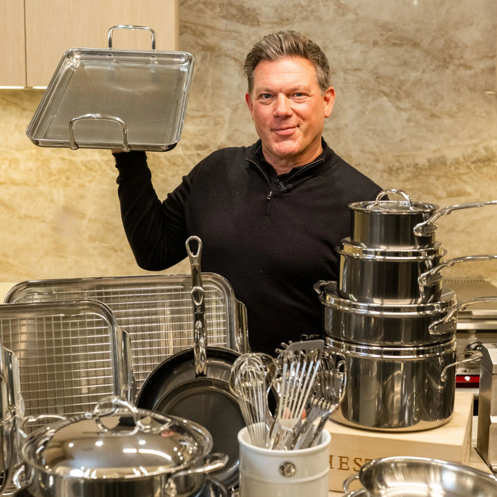 A man in a black shirt stands in a kitchen, holding up a metal baking tray. In front of him are various stainless steel pots, pans, lids, and whisks from the NanoBond® Titanium x Tyler Florence Cookware Set, 41-Piece—each piece neatly organized on the counter and celebrated for their distinctive Molecular Titanium finish.