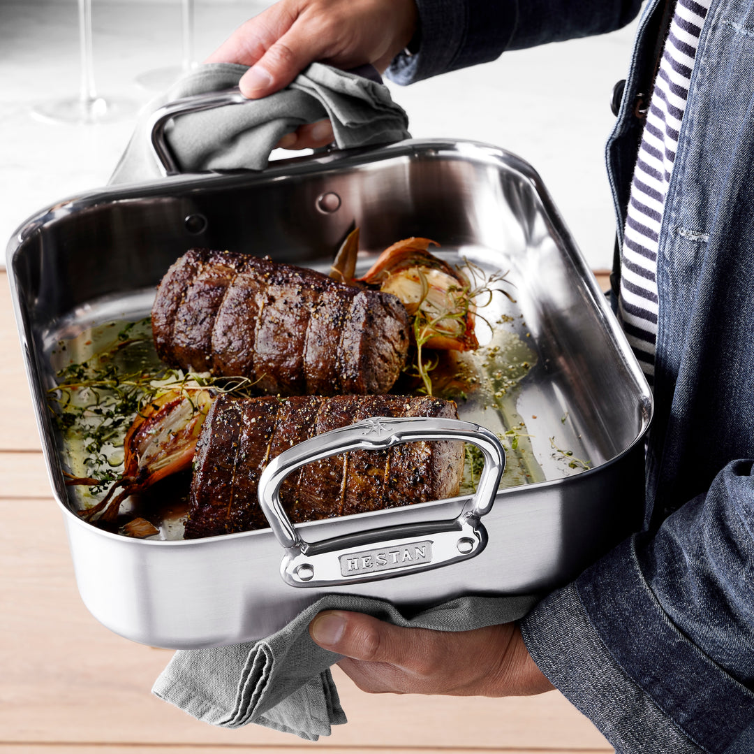 A person holds a stainless steel roasting pan with two roasted beef tenderloins seasoned with herbs. The pan rests on a table, and the person wears a striped shirt and denim jacket. A towel is used to hold the pan handles, and the brand Hestan is visible.