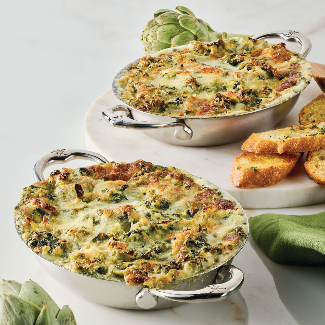 Two oval metal dishes filled with baked spinach artichoke dip are placed on a marble surface. Each dish is accompanied by slices of toasted bread. A green napkin and an artichoke are visible nearby.